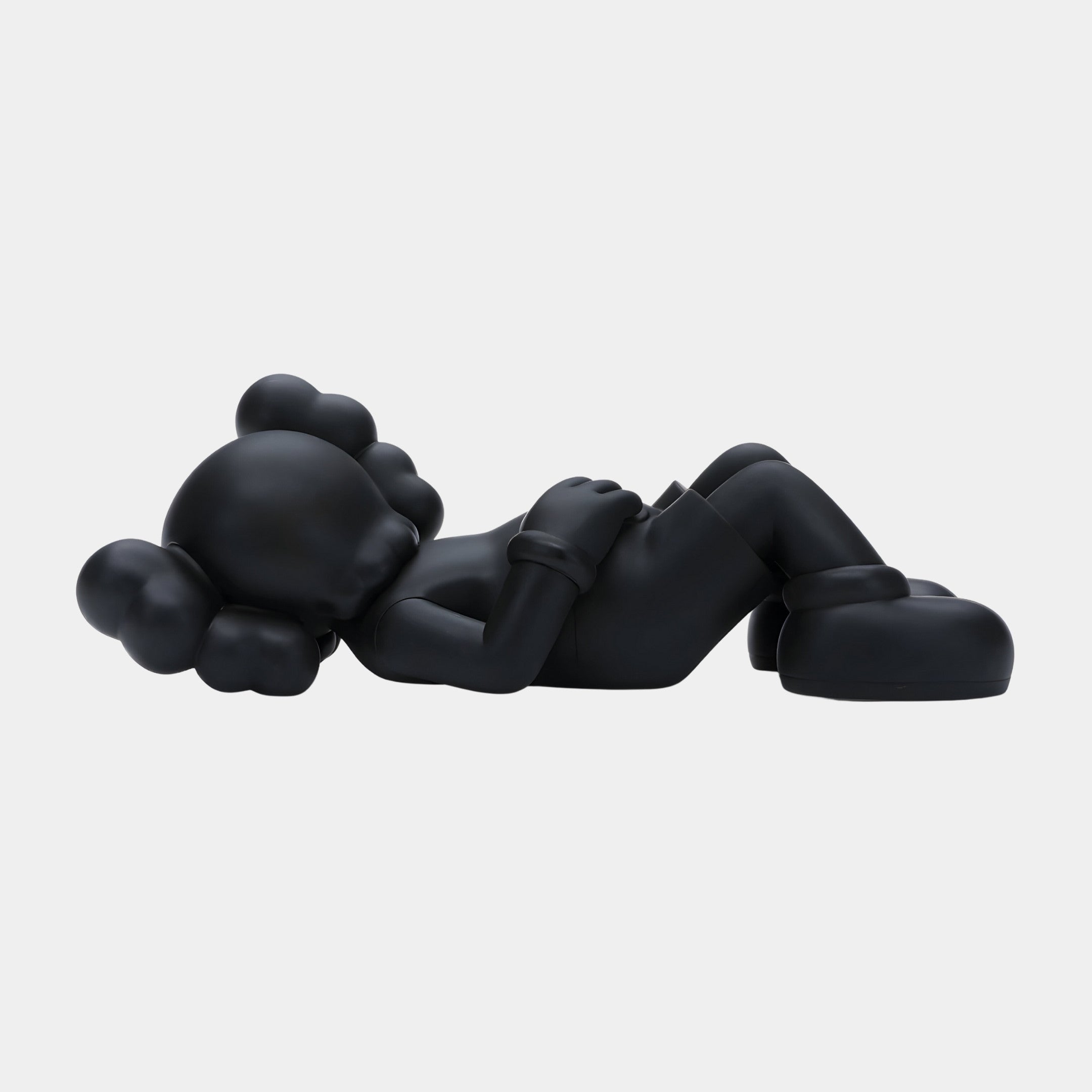 The 110cm Obsidian Black Iconify Peace Sculpture from Giant Sculptures features a cartoonish figure lying on its back with round, bulbous features, hands on its chest, knees bent, and wearing gloves and shoes. It has a smooth matte finish that enhances its modern aesthetic against a plain white background.