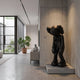 Midnight Black Iconify Burden Sculpture, 130cm, standing in a modern hallway on a concrete plinth against a textured wall, near a potted plant and console table.