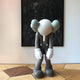 A hazelnut brown cartoonish figure with a round head and large hands, resembling the Giant Sculptures Iconify Burden Sculpture, stands with its head bowed and arms hanging like an elephant with two big ears, no facial features. A dark abstract painting adorns the wall behind it.