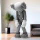 A 130cm Slate Grey Iconify Burden Sculpture features a cartoonish figure with crossed-out eyes and X-shaped hands by Giant Sculptures. Its bowed head and large ears convey introspection in a modern room, complemented by a potted plant and sofa for serene elegance.