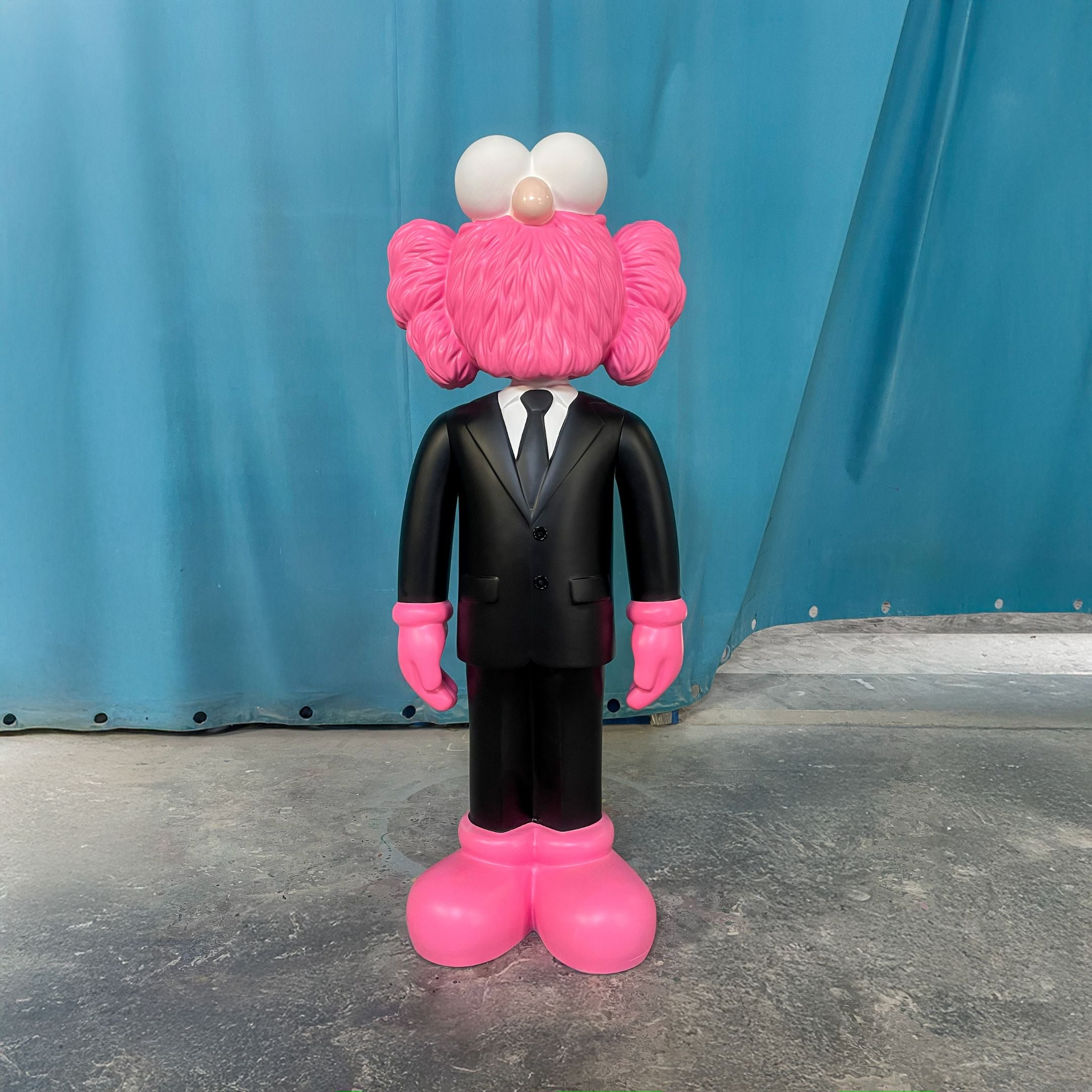 The Black Suit Iconify CEO Sculpture - 130cm by Giant Sculptures features a pink, furry figure with cartoonish details and oversized hands and feet, standing on concrete before a blue curtain—a bold resin piece for luxury interiors.