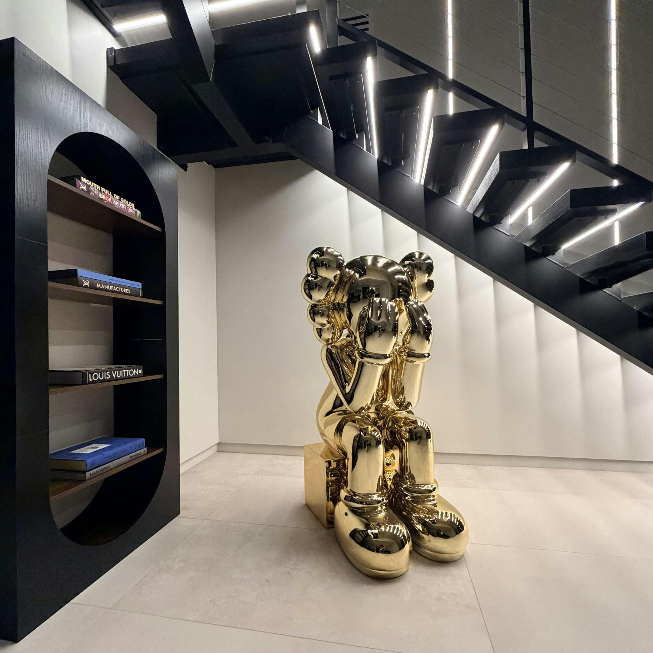 The Antique Gold Iconify Melancholy Sitting Sculpture (130cm) by Giant Sculptures features a cartoon-like figure with its head in its hands, sitting in gold beneath a black staircase; nearby, a black shelf displays luxury-brand books.