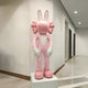 The Giant Sculptures Coral Pink Iconify Rabbit Sculpture - 130cm stands on a white pedestal in a modern hallway. Its large ears, puffy cheeks, and oversized hands and feet complement the stylish living rooms contemporary design, harmonizing with the white walls and beige flooring.