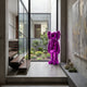 Phantom Purple Iconify Standing Sculpture - 135cm, glossy metallic-purple figure standing by a glass wall in a modern hallway with a wood staircase and courtyard view.