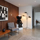 Obsidian Black Iconify Furry Sitting Sculpture, 140cm, seated on white platform in living room beside grey chair, wood-slat wall, and black tripod floor lamp.