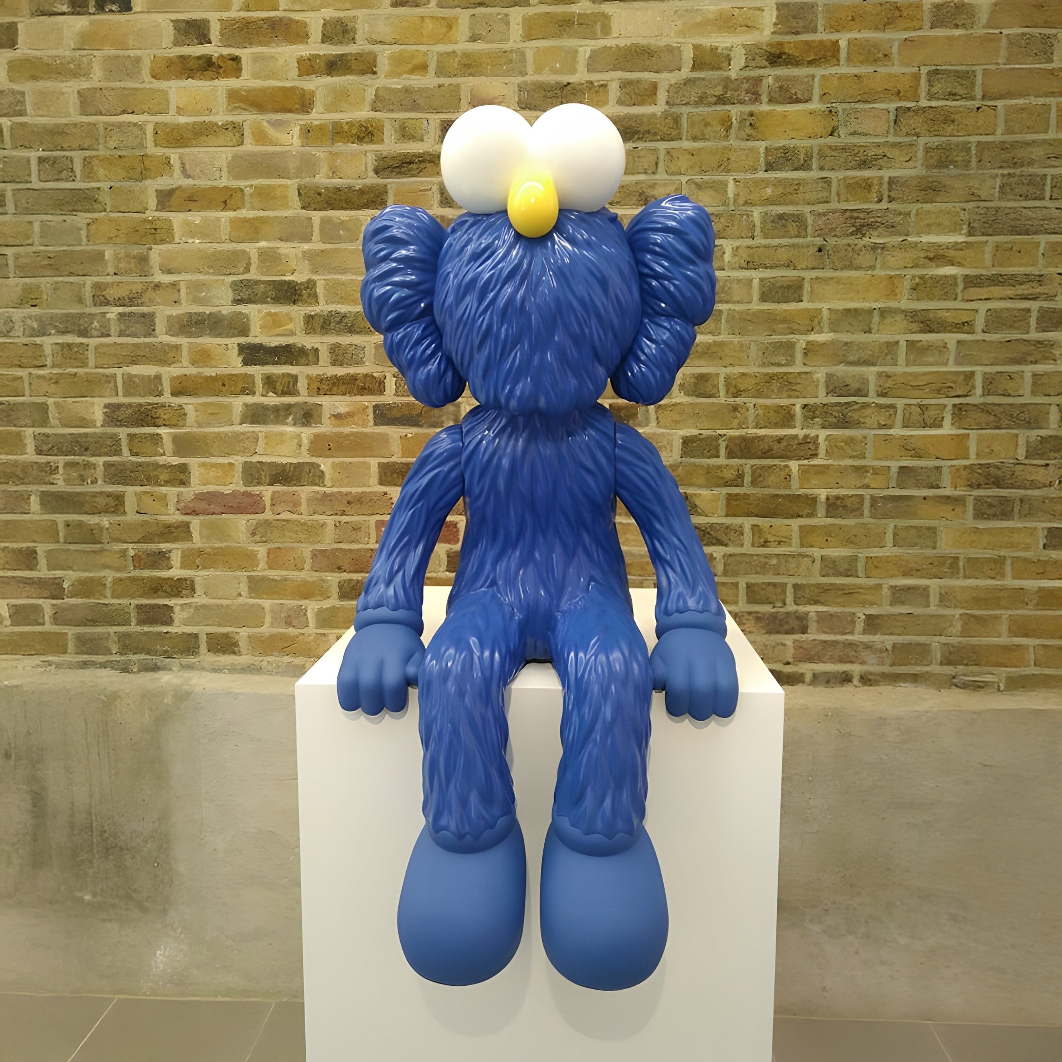 The Deep Blue Iconify Furry Sitting Sculpture by Giant Sculptures, measuring 140cm with oversized eyes and long ears, rests on a white pedestal. Its fuzzy texture and vibrant blue color pop vividly against the brick wall backdrop, capturing the essence of contemporary pop art.