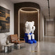 White & Blue Iconify Melancholy Sitting Sculpture, 163cm resin figure with head in hands, shown in a wood-paneled hallway niche beside a leather armchair.