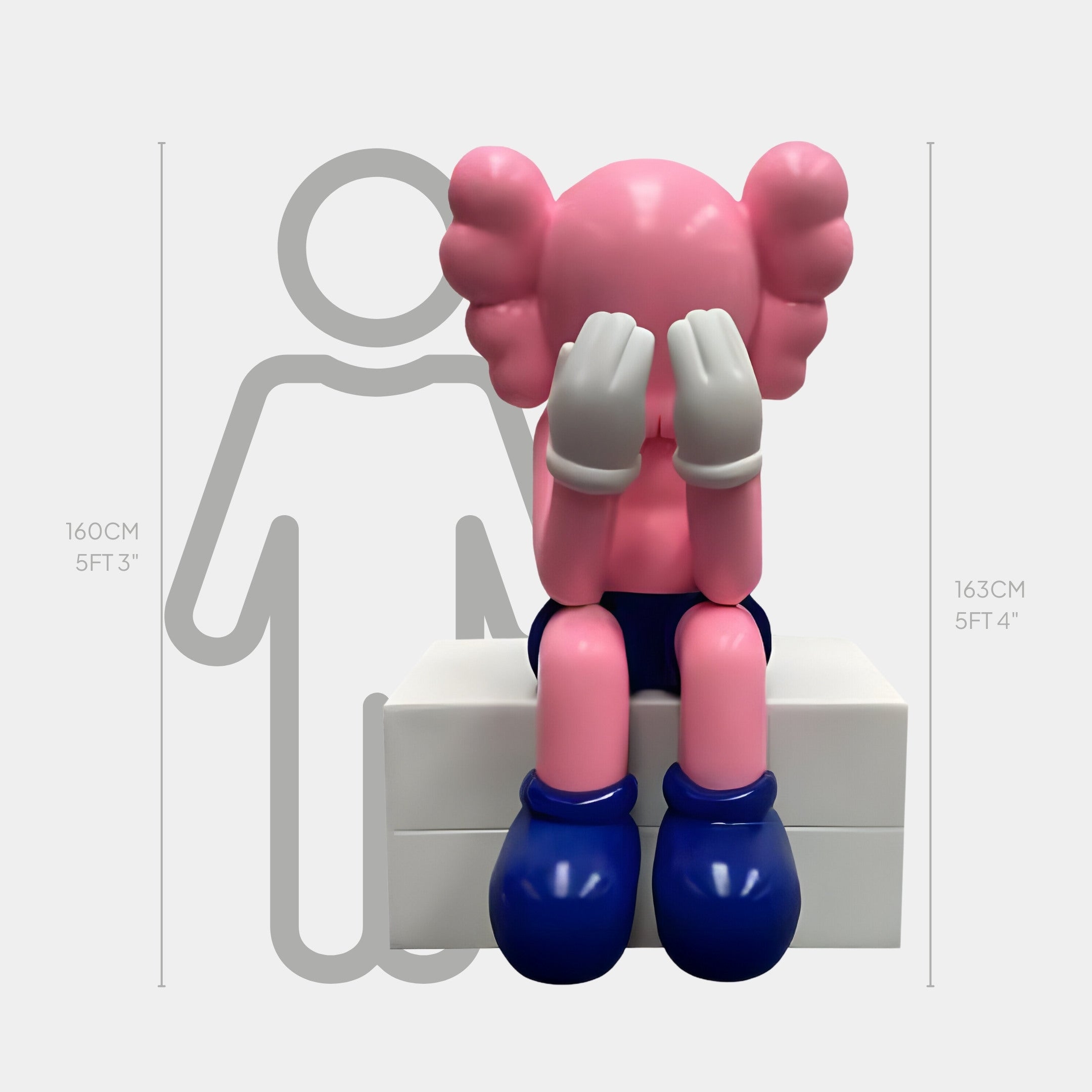 The Petal Pink Iconify Melancholy Sitting Sculpture by Giant Sculptures features a cartoon-like figure with large blue shoes and gloved hands covering its face, perched on a white block. A gray silhouette with an emotional expression complements this modern piece, marked at 5 ft 3 in and 5 ft 4 in.