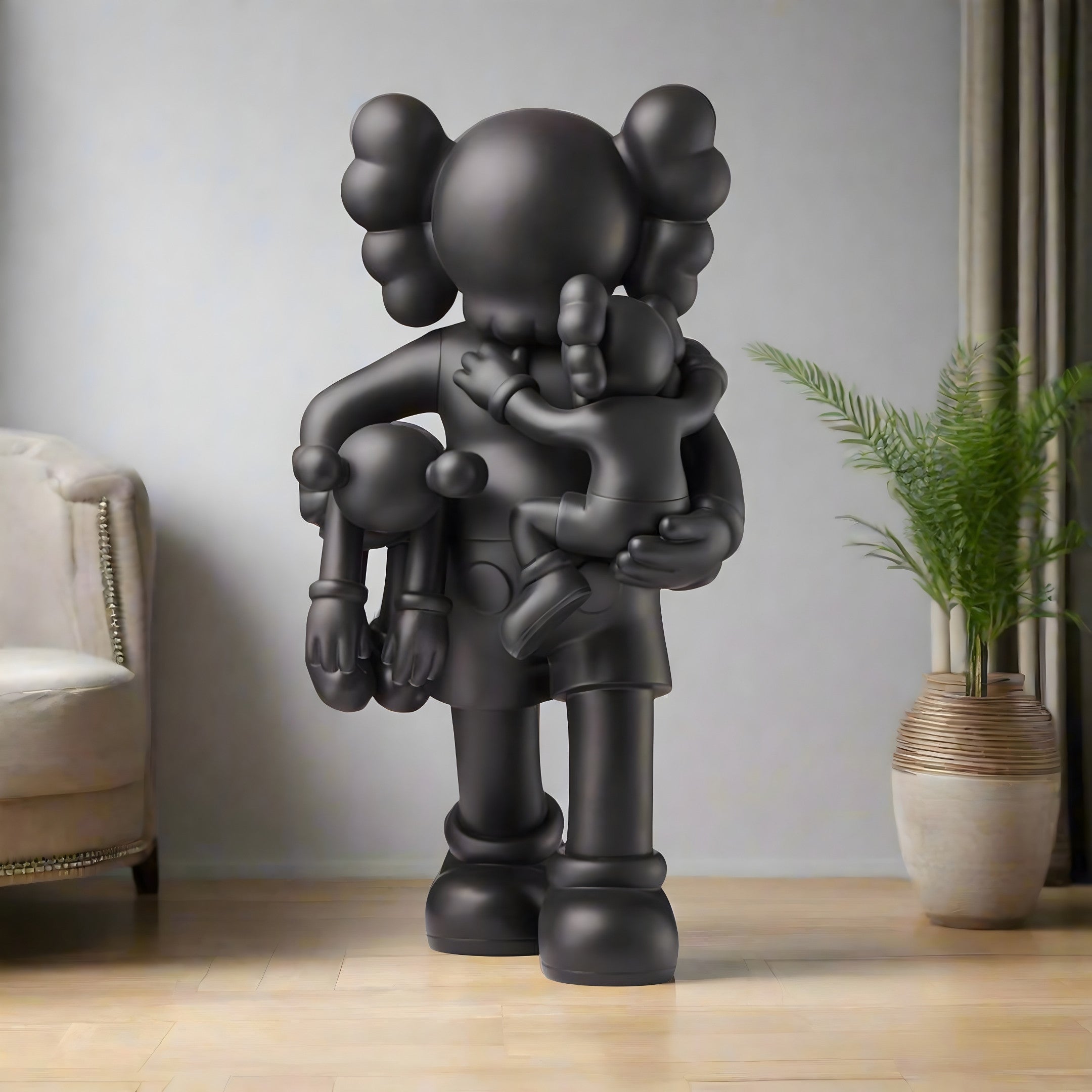 The 180cm Midnight Black Iconify Family Sculpture by Giant Sculptures depicts a dark gray, round-headed figure with glove-like hands holding two smaller figures. Positioned in a room with a light sofa, curtains, and leafy potted plant on a wooden floor, it adds modern artistic expression.