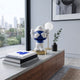 Deep Blue Iconify Peekaboo Figure, 20cm, covering its face with white hands on a wooden console beside books, a globe lamp, and framed art.
