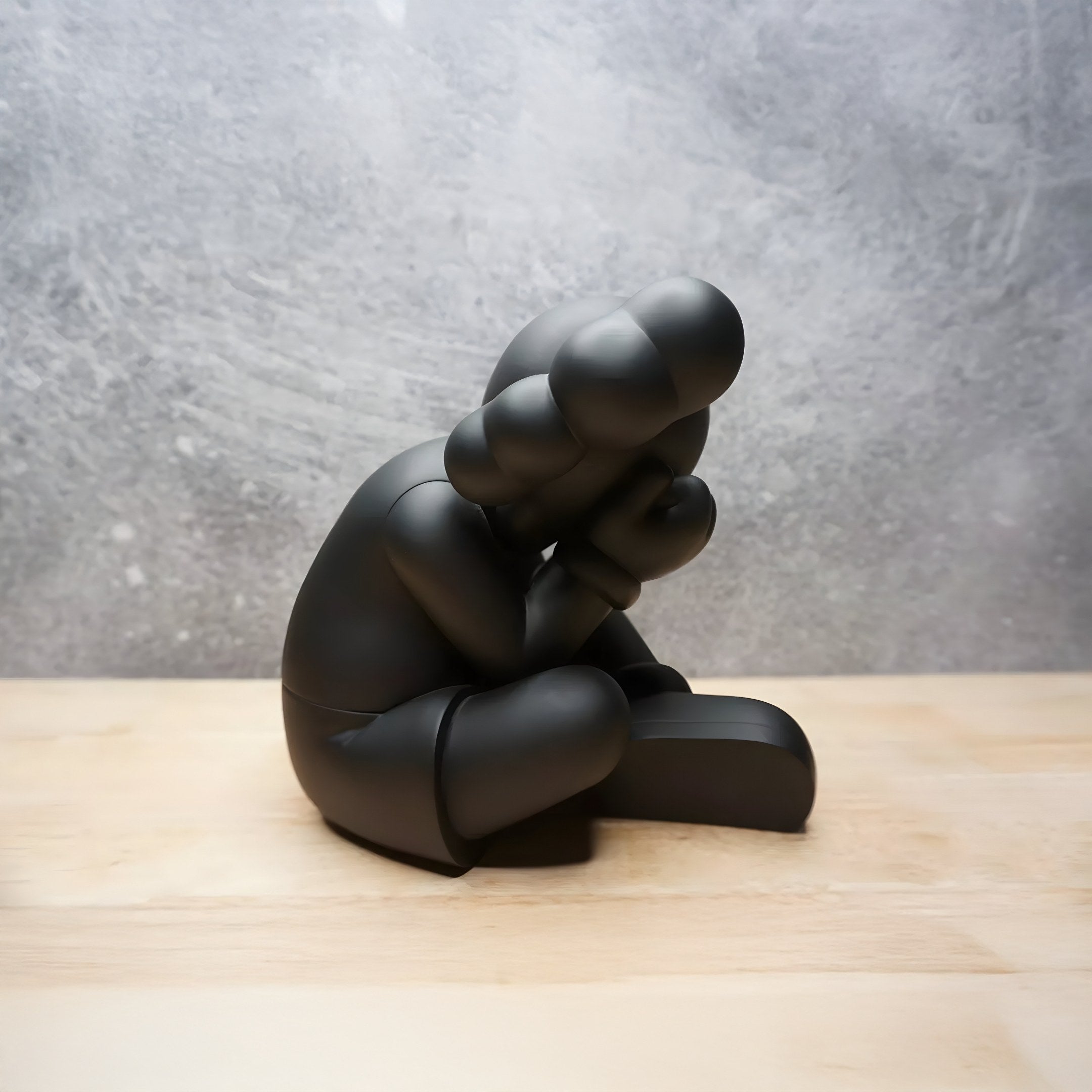 The Giant Sculptures Midnight Black Iconify Sorrow Figure - 20cm depicts a stylized form with head on knees and arms over its face. Its rounded shapes intrigue as it sits on wood against a textured gray backdrop, ideal for contemporary art displays.