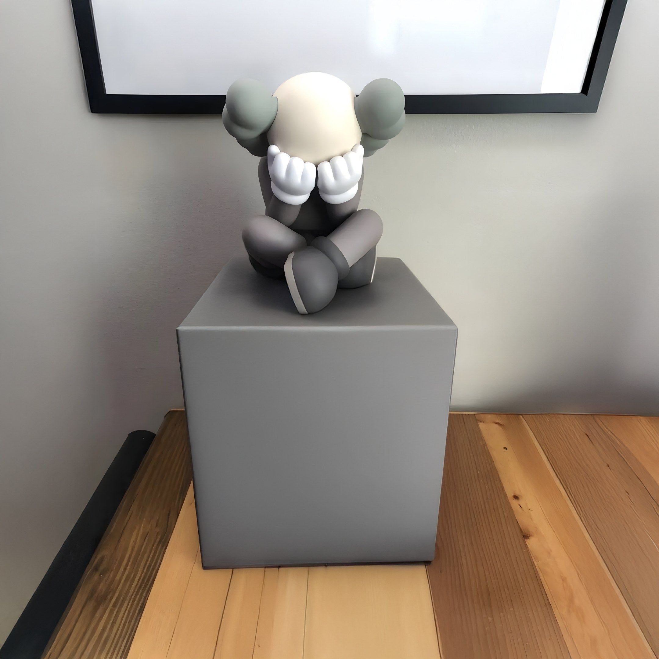 The Hazelnut Brown Iconify Sorrow Figure - 20cm by Giant Sculptures features a stylized character seated on a gray pedestal, face hidden by gloved hands. Set indoors with a wooden floor and blank framed canvas behind it, this piece beautifully embodies contemplation.