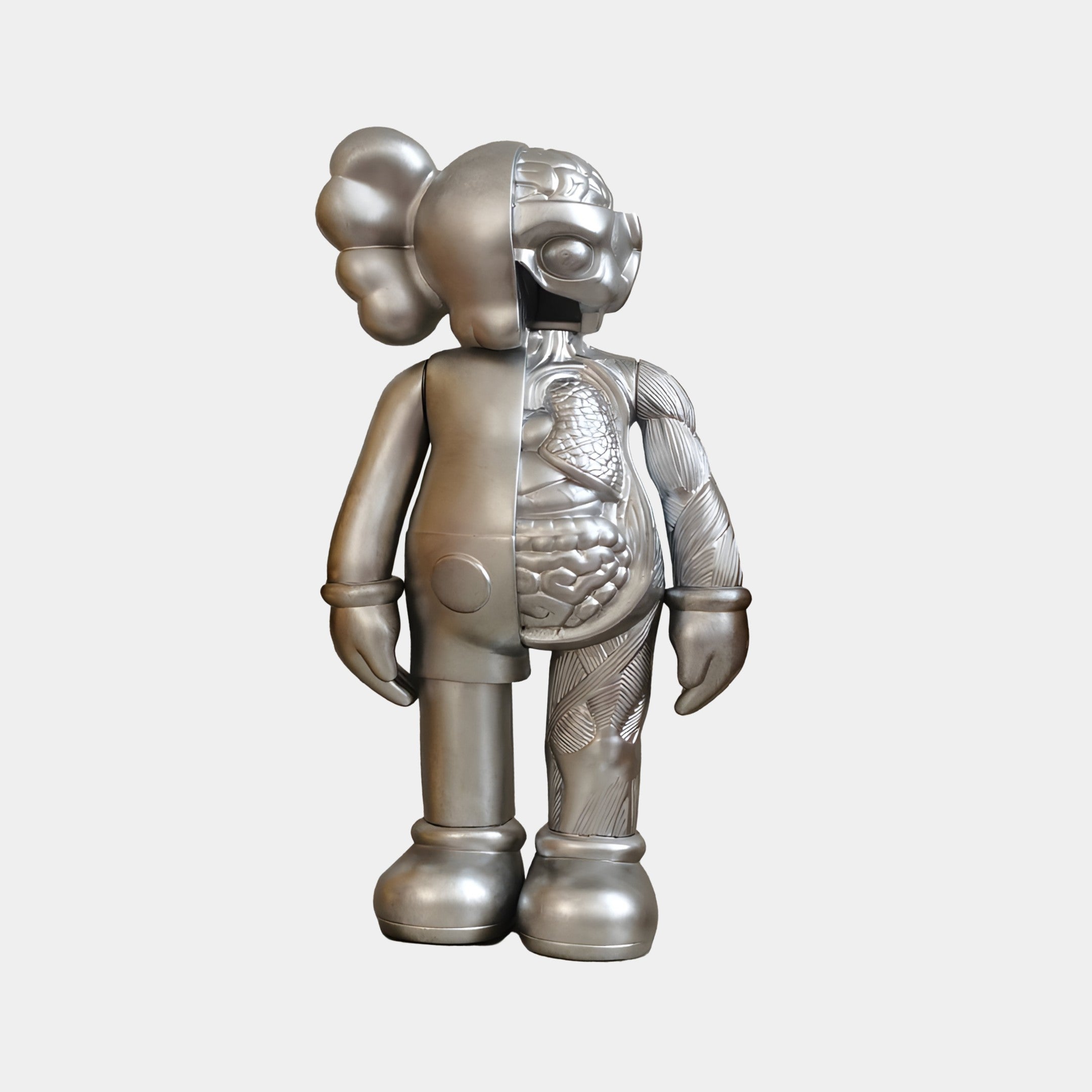 The Titanium Slate Iconify Anatomy Figure by Giant Sculptures is a 20cm silver sculpture of a standing cartoonish figure with a large head, featuring one half with intricate internal structures and the other smooth. It combines anatomical artistry with whimsy, sporting large gloves and shoes.