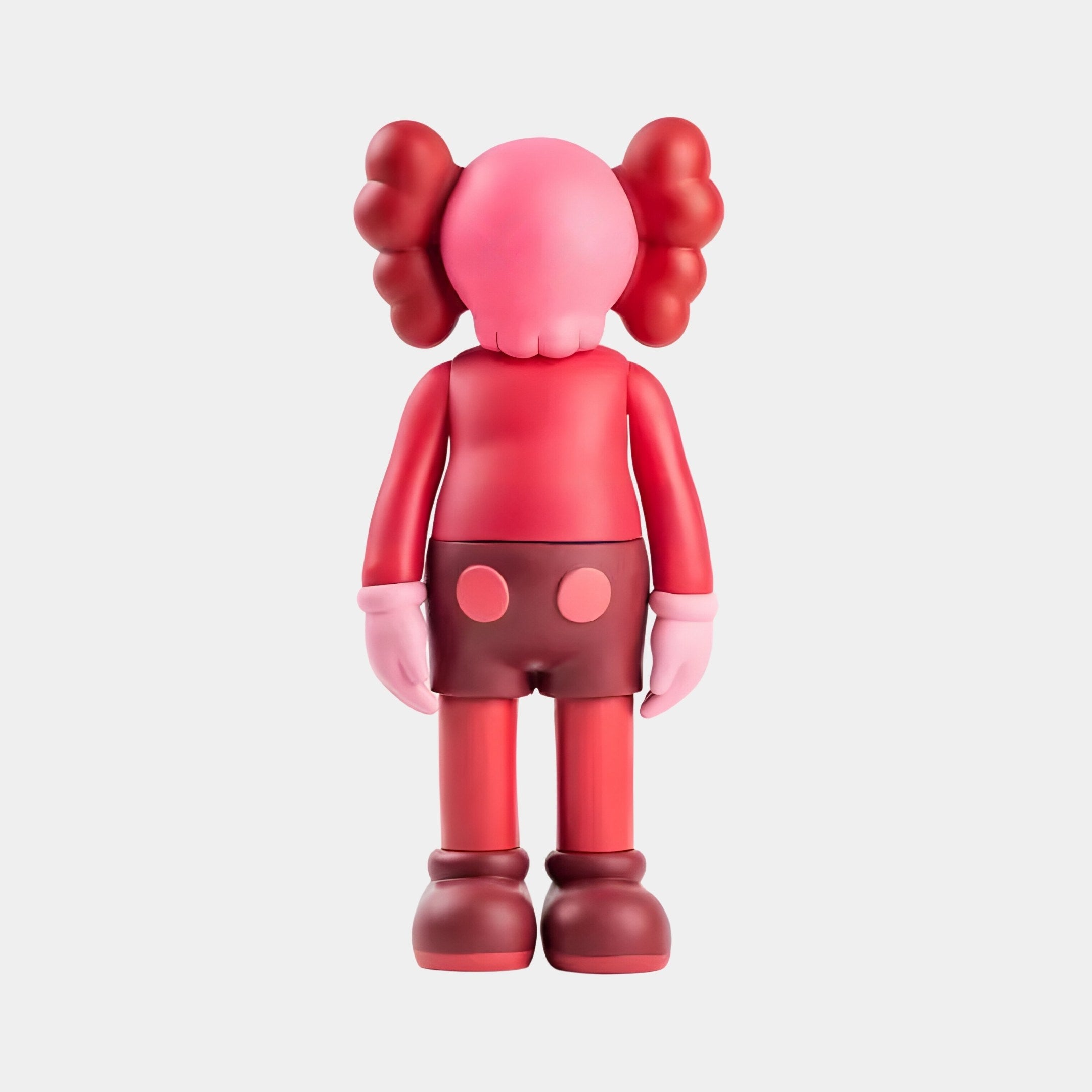 The Blush Ruby Iconify Standing Figure - 20cm by Giant Sculptures exudes energetic charm with its vibrant vinyl design featuring a pink head and hands, red shirt, brown shorts, cloud-like red shapes, and brown shoes reminiscent of an exquisite resin sculpture.