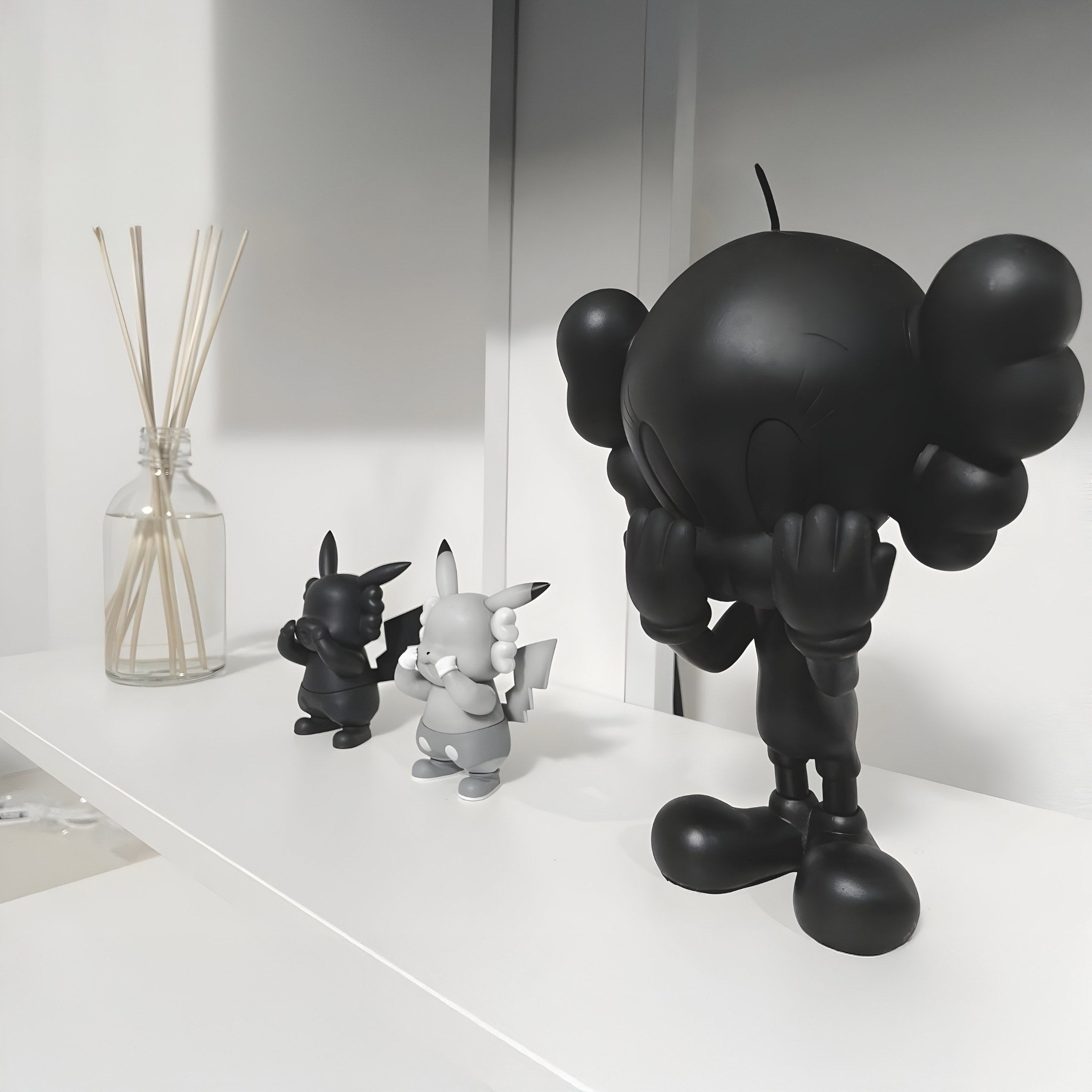 The Black Iconify Tweeto Figure (20cm) by Giant Sculptures stands prominently on a white shelf with its hands on its face, flanked by two smaller black-and-white abstract figurines. A glass bottle with reed sticks adds a finishing touch to this monochrome display.