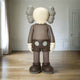 The Hazelnut Brown Iconify Standing Sculpture by Giant Sculptures is a large, cartoon-like figure crafted from premium resin, featuring a round head with X eyes, grey squiggly ears, and wearing white gloves and shoes paired with hazelnut brown shorts. It stands in an empty room with wooden floors and white walls.