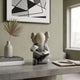 Soft Grey Iconify Hollow Figure - 23cm, seated cross-legged with skull-like head and white gloves, displayed on a stone table beneath framed abstract wall art.