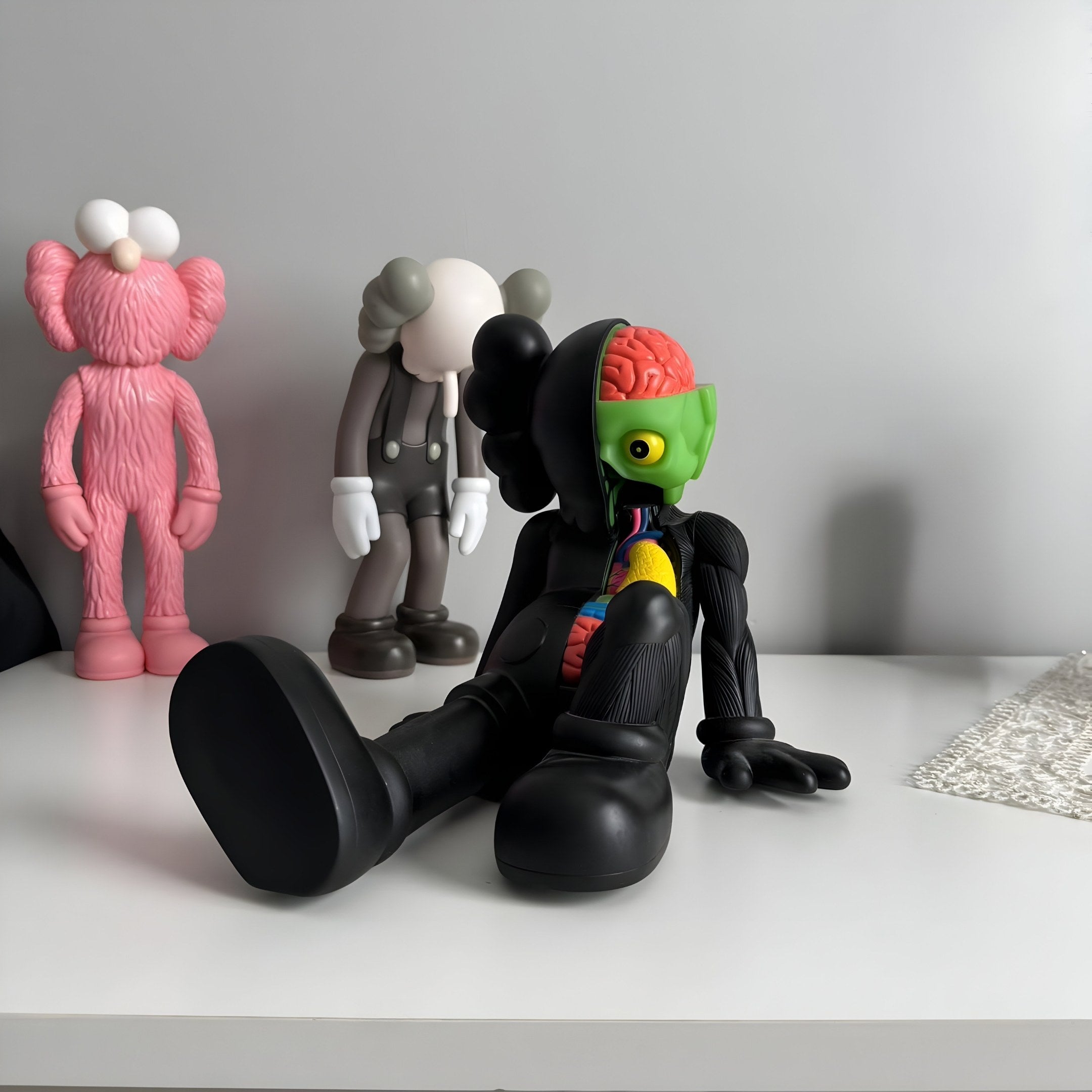 On a white surface sits the Midnight Black Iconify Anatomy Figure - 23cm by Giant Sculptures, showcasing colorful anatomical details like a green head with a visible brain. Behind it are two quirky characters with distinct pink and gray features.