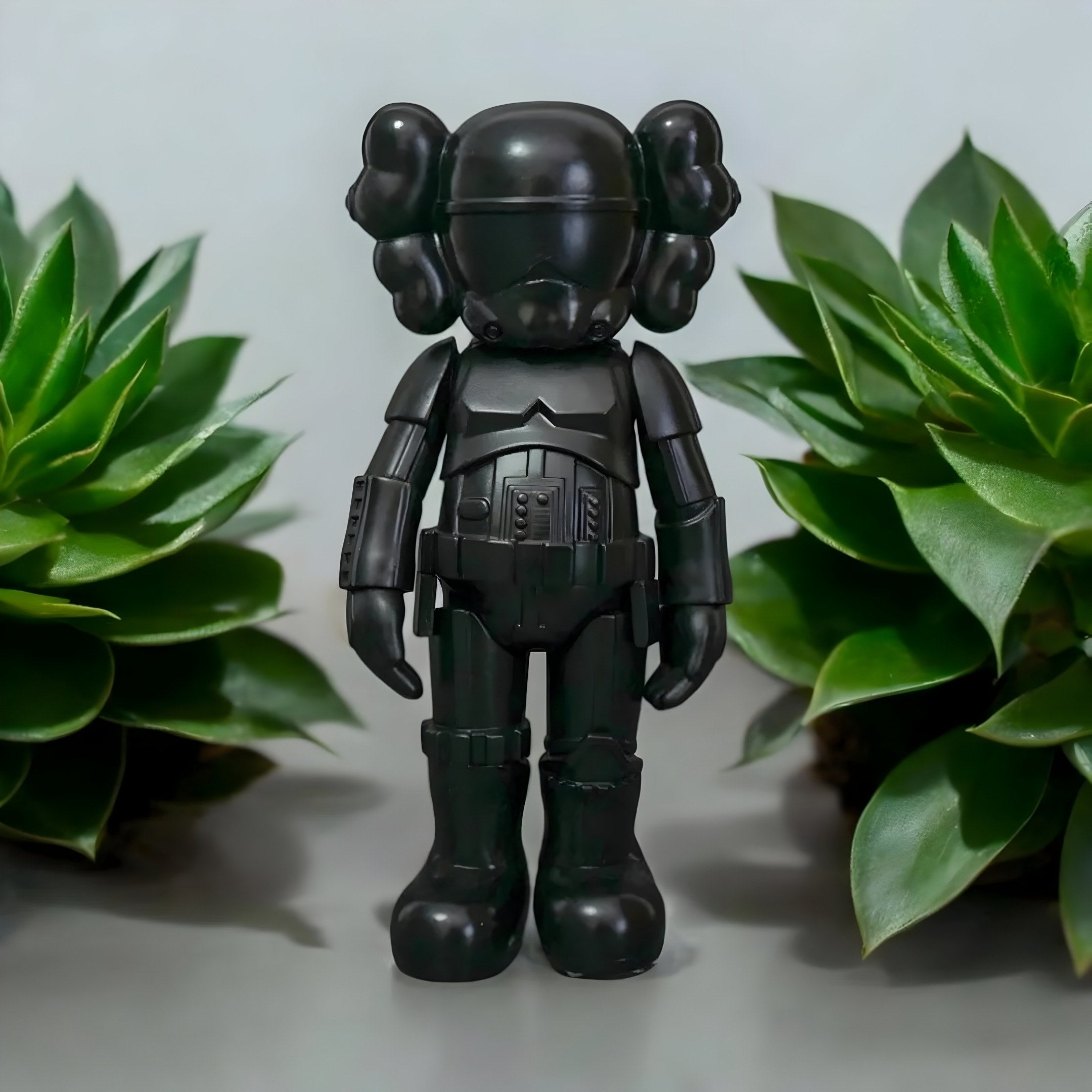 The Midnight Black Iconify Enforcer Figure by Giant Sculptures is a 26cm black resin figure with a helmet and exaggerated features, flanked by green succulents on a gray background—ideal for modern interiors.