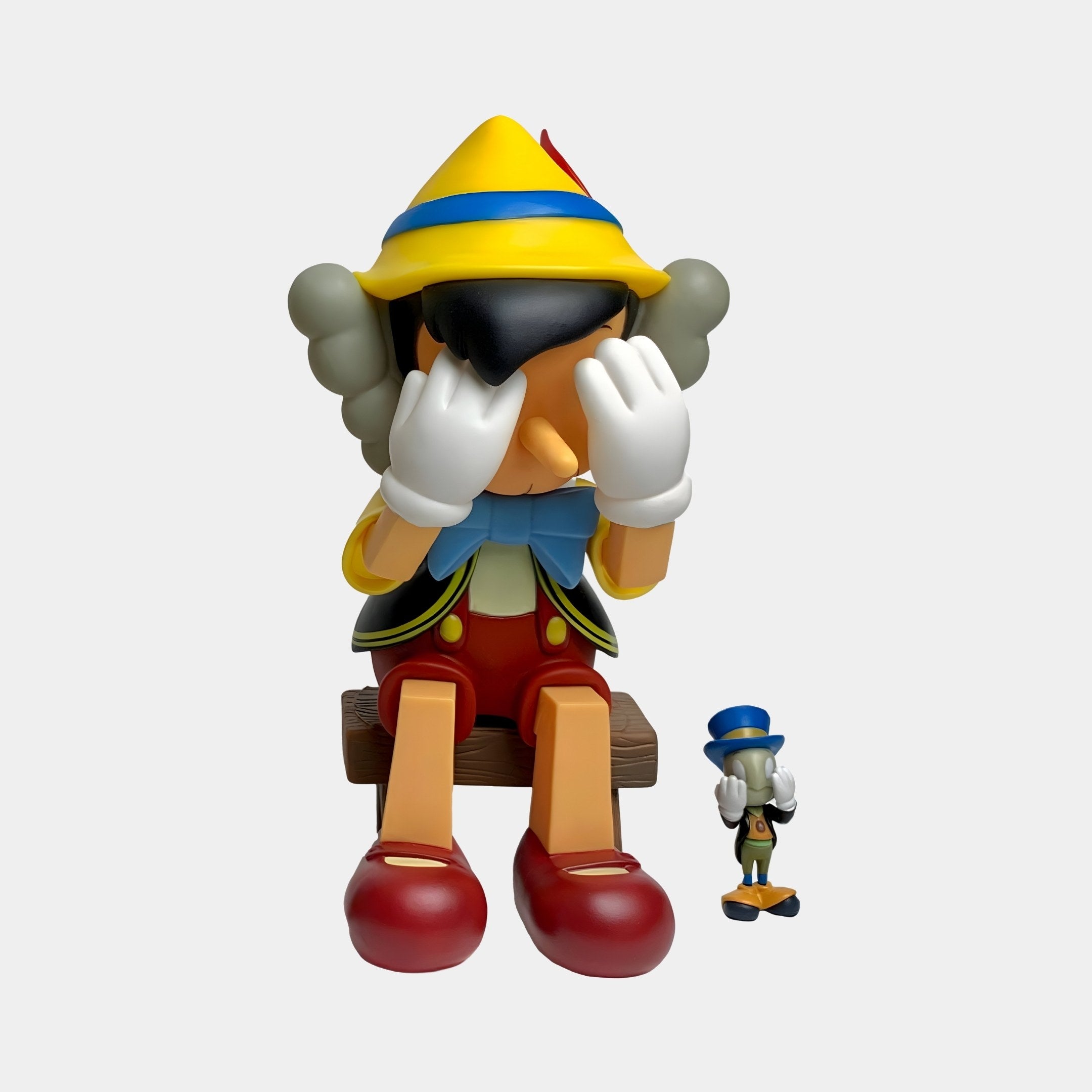 A pop-art Iconify Marionette Sitting Figure by Giant Sculptures, measuring 26cm, sits on a wooden bench with gloved hands covering its eyes. Next to it, a whimsical storytelling cricket sculpture in a top hat looks up at the larger iconified figure.