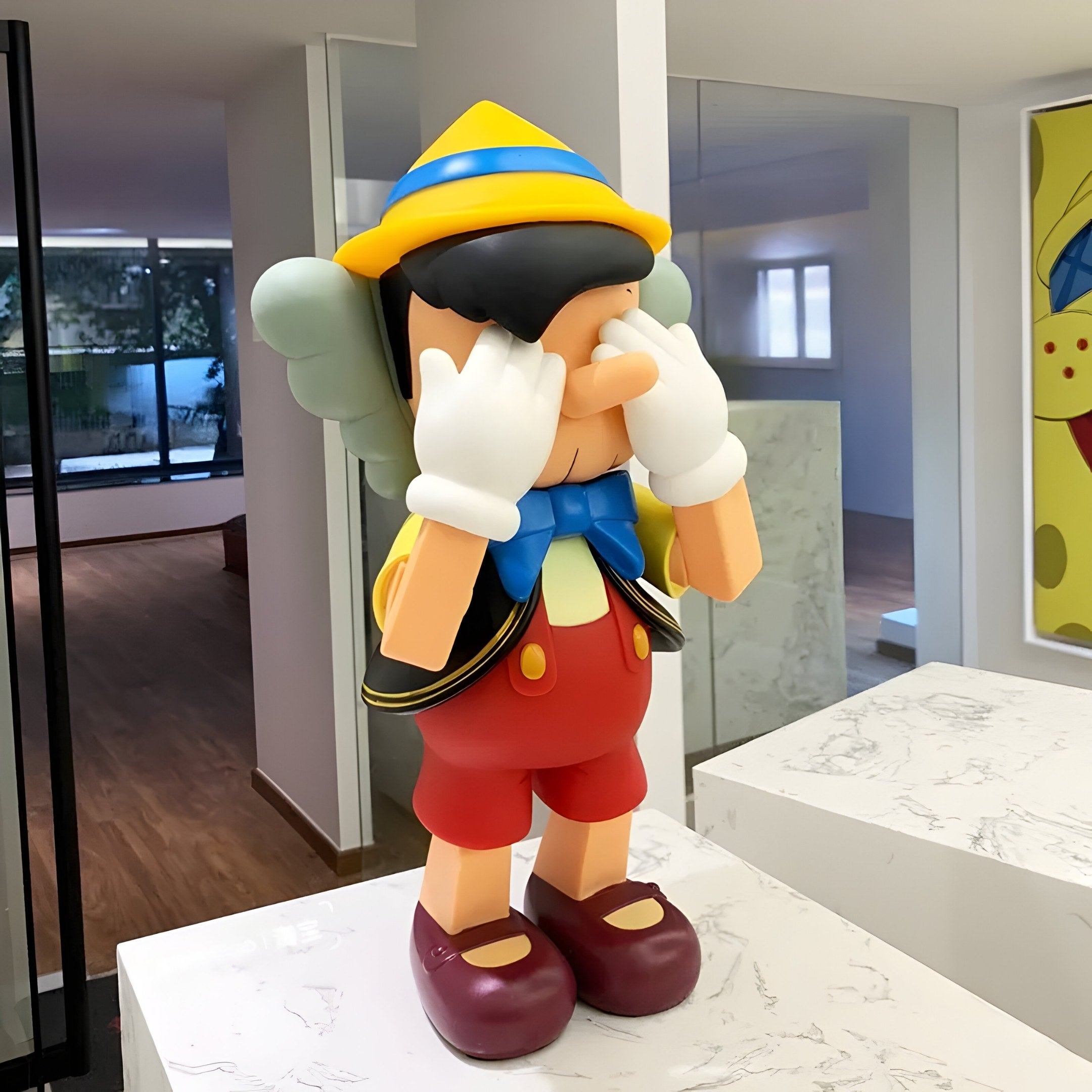 In a modern room, the Iconify Marionette Figure by Giant Sculptures captivates—this 43cm premium resin piece depicts a cartoon character with long nose, red shorts, white gloves, yellow hat, and blue bow tie covering its eyes with hands on a sleek marble surface.