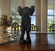 A Giant Sculptures Midnight Black Iconify Burden Figure - 28cm, with X-marked eyes, sits on a polished wooden table next to a shiny silver trophy, oozing pop-art charm. Large windows behind frame views of trees and sky.