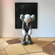 A Hazelnut Brown Iconify Burden Figure by Giant Sculptures leans with its oversized head echoing pop-art influences, slouched in a room with wooden floors and set against a dark abstract painting backdrop.