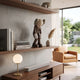 Hazelnut Brown Iconify Burden Figure, 28cm, displayed on a wooden wall shelf beside books and a vase in a living room with a tan armchair.
