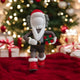 The 28cm Iconify Christmas Figure by Giant Sculptures features a cartoon Santa holding a green and red gift, set on red cloth against a backdrop of modern holiday décor with a decorated tree and blurred presents.