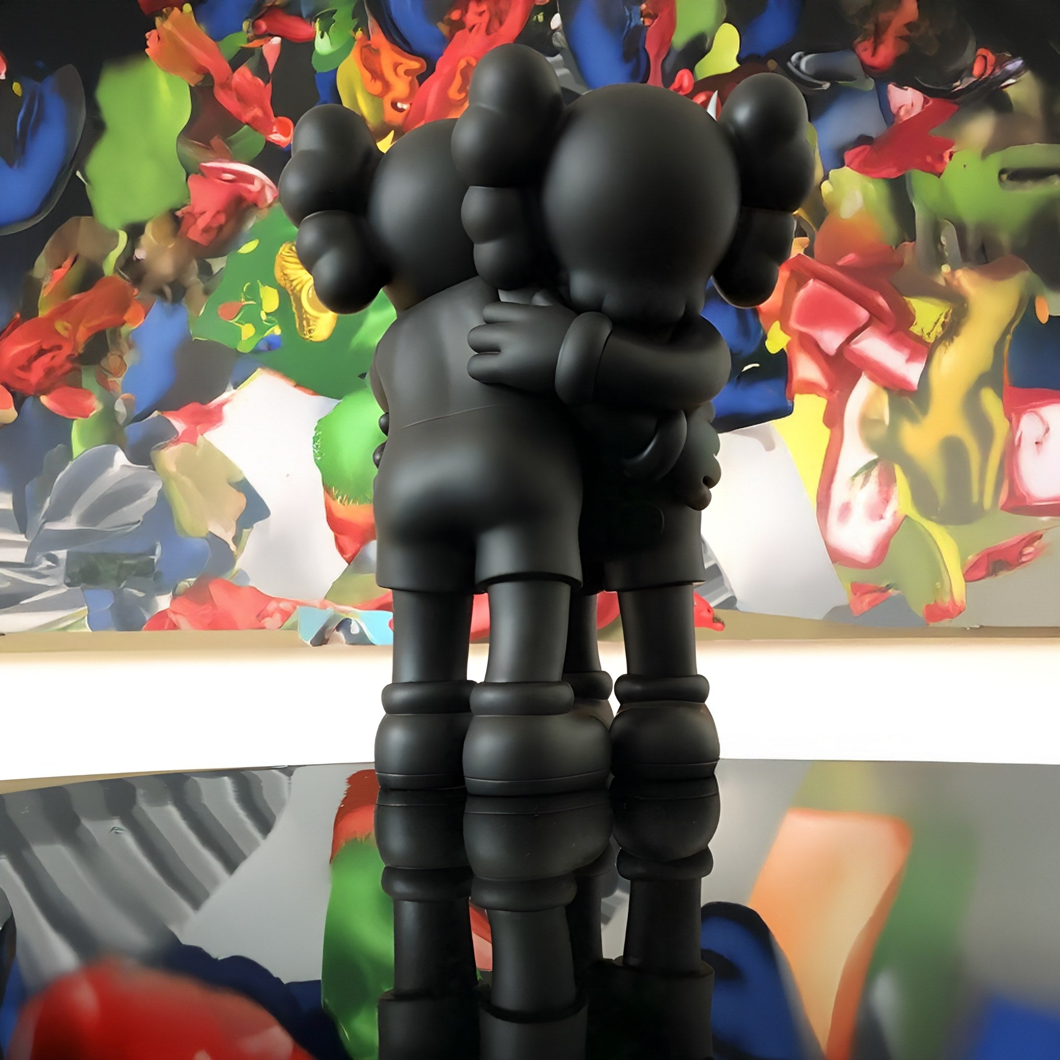 On a reflective surface, the Giant Sculptures Obsidian Black Iconify Embrace Figures—28cm tall with rounded shapes—stand united. Set against a colorful, abstract backdrop of various forms and hues, this resin sculpture captures the essence of human connection.