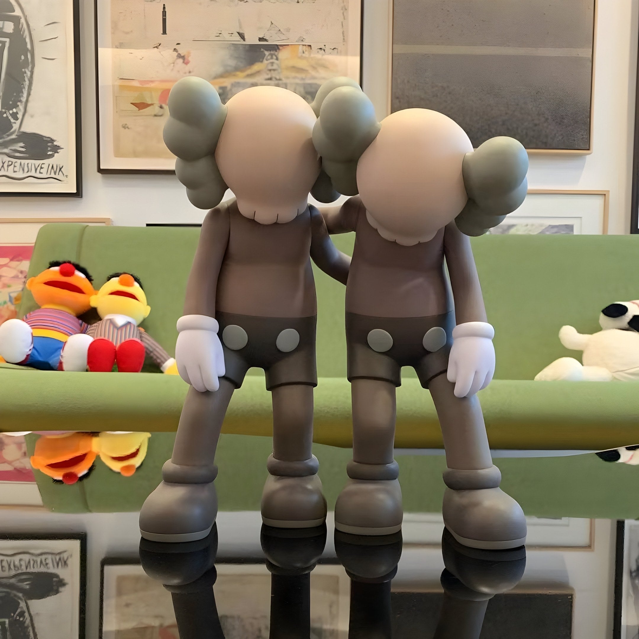 Two Hazelnut Brown Iconify Friendship Figures by Giant Sculptures, featuring large hands and crossed-out eyes, stand on a reflective surface. Behind them are plush toys with orange and yellow features on a green couch in front of framed art.