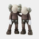 The Hazelnut Brown Iconify Friendship Figure from Giant Sculptures features two gray, cartoon-like figures with large heads and crossed-out eyes. They stand side by side in outfits with large buttons, gloved hands, and one arm around each others shoulder. Height: 28cm.