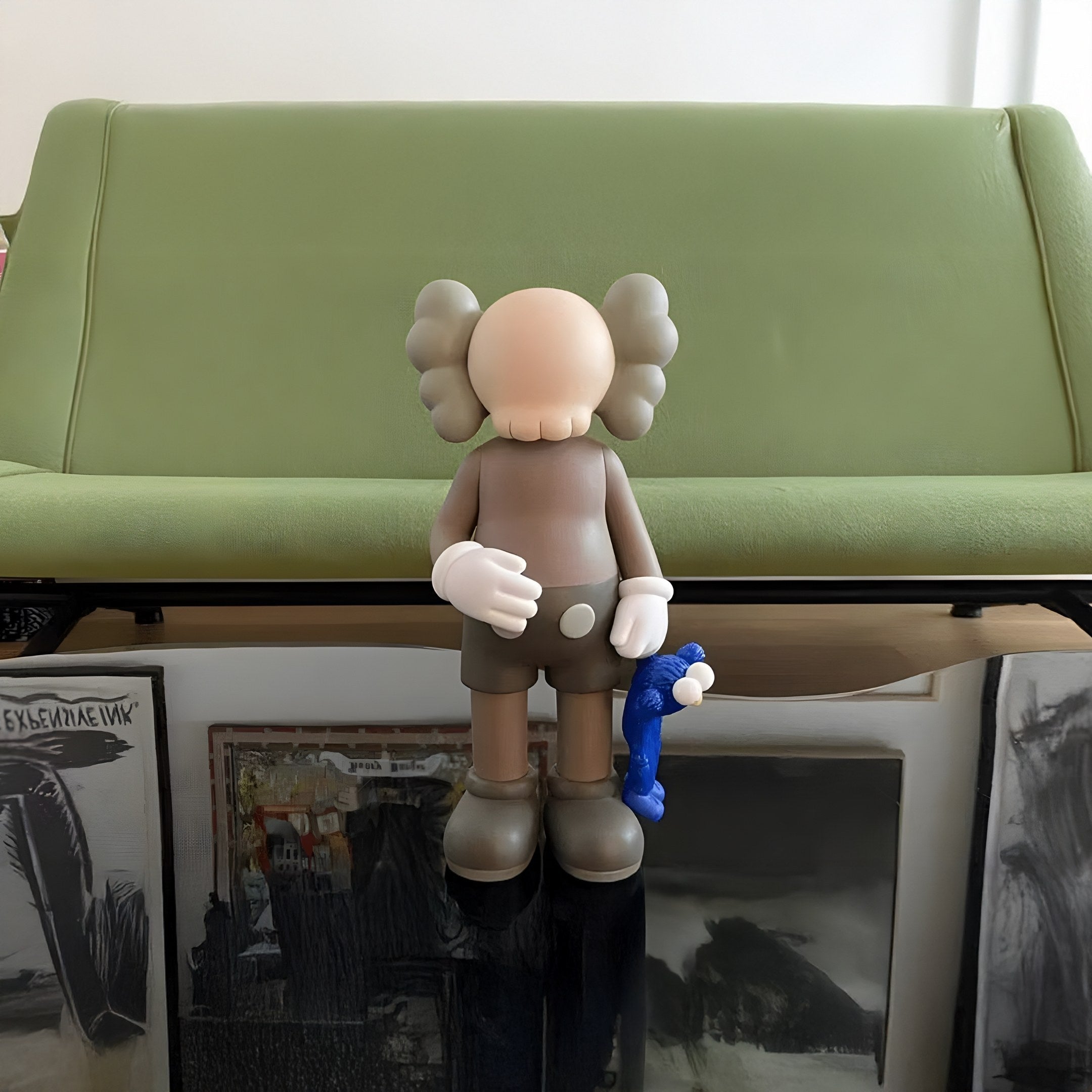 The 28cm Hazelnut Brown Iconify Blue Furry Toy Figure by Giant Sculptures stands on a reflective surface in front of a light green sofa, grasping a small blue item. The background features various artworks leaning against the wall, making it a true statement piece.