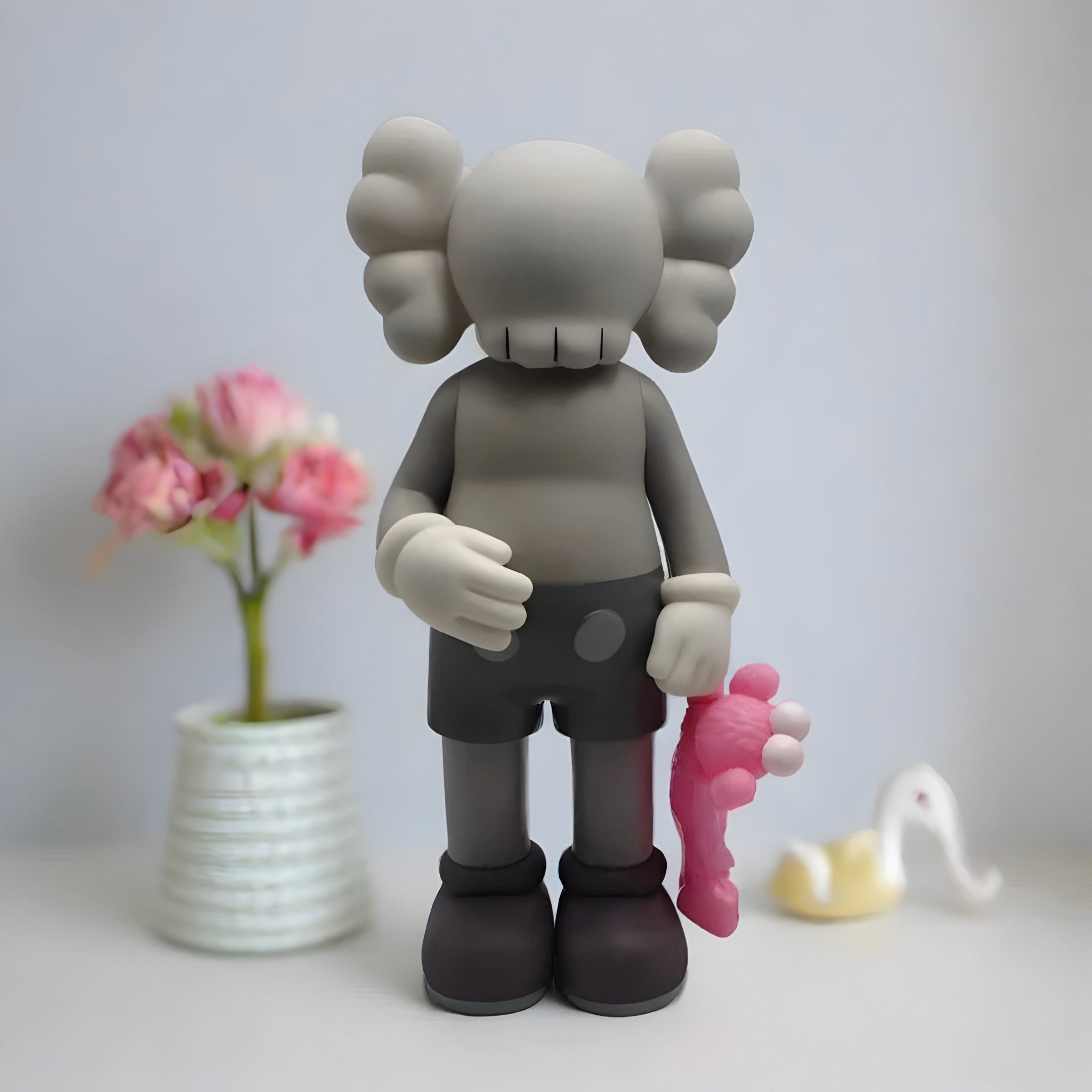 The 28cm Soft Grey Iconify Pink Furry Toy Figure by Giant Sculptures features a gray, cartoonish figure with large ears and X-eyes, holding a small pink object. A vase with pink flowers and an abstract white piece in the background add to the modern sculpture vibe.