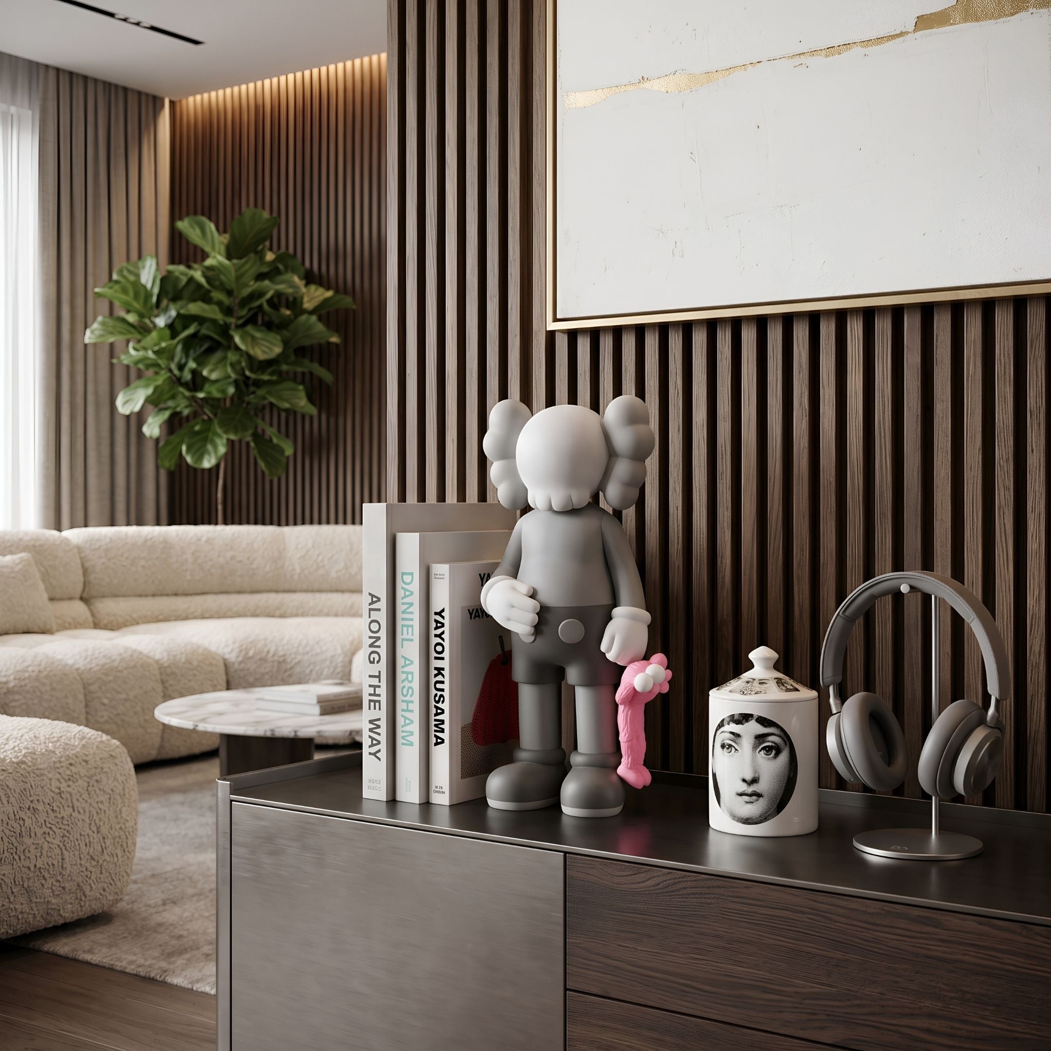 Soft Grey Iconify Pink Furry Toy Figure, 28cm, by Giant Sculptures, shown on a dark sideboard beside books, a headphone stand, and a printed jar in a living room setting.