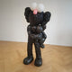 The Giant Sculptures Iconify Loving Furry Figure is a large, obsidian black sculpture with oversized eyes and a pink nose on a light wooden floor. It embraces a smaller figure in a playful, cartoonish style, showcasing pop-art flair. Length: 28cm.