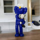 The Giant Sculptures Deep Blue Iconify Loving Furry Figure - 28cm, a deep blue furry cartoon character with large white eyes, embraces a smaller gray companion on a wooden surface against contemporary interiors with shelves and light-colored walls.