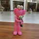 The Petal Pink Iconify Loving Furry Figure, a 28cm resin sculpture by Giant Sculptures, stands on an indoor wooden floor. With its large eyes, it lovingly cradles a small gray figure against a backdrop of blurred interior elements.