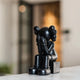 The Giant Sculptures Midnight Black Iconify Melancholy Sitting Figure - 28cm is a resin sculpture of a figure sitting with gloved hands over its face, resting on a square block. Its spherical head has ear-like features and is set against a softly blurred indoor backdrop.