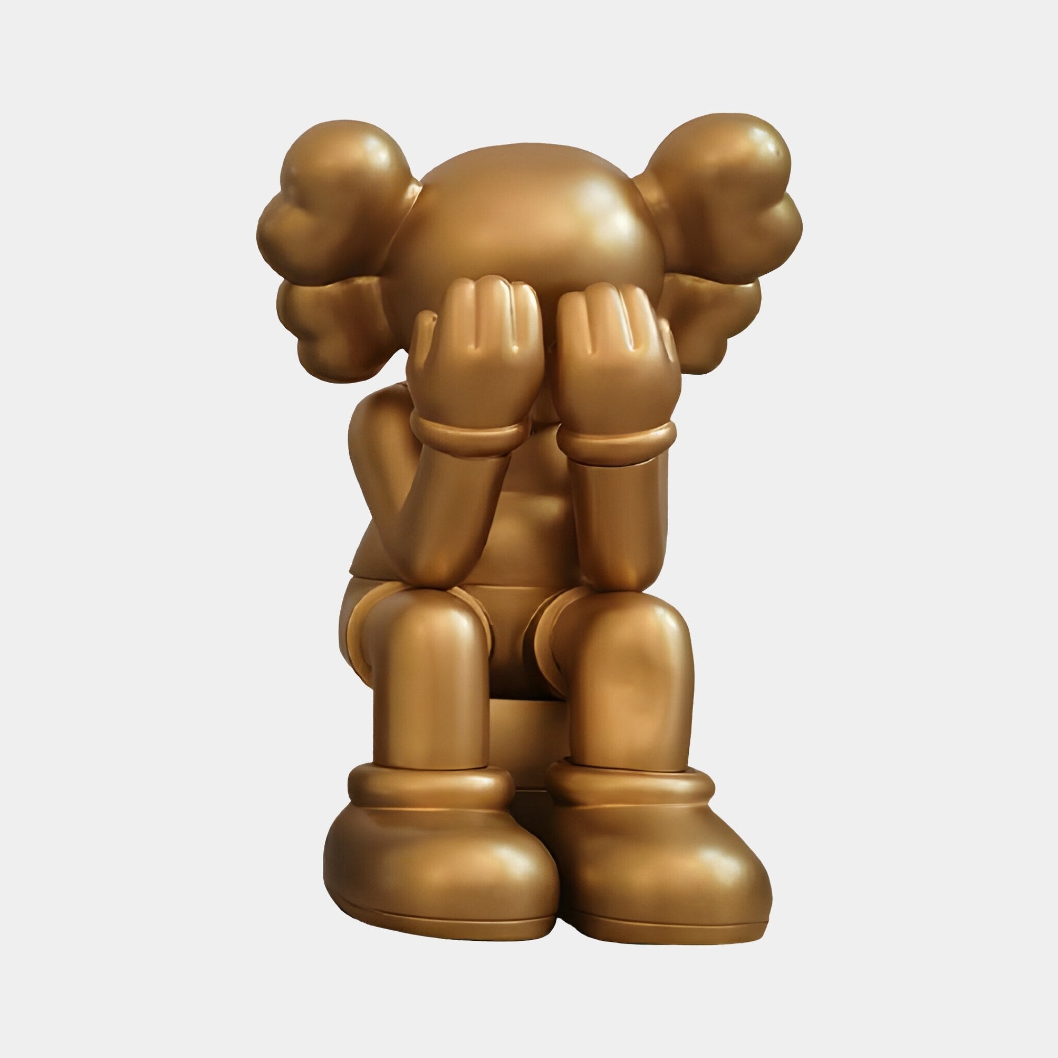 The Champagne Satin Gold Iconify Melancholy Sitting Figure by Giant Sculptures is a 28cm resin sculpture featuring a stylized figure with bulbous, rounded features, sitting with hands covering its face in a thoughtful or melancholy pose.