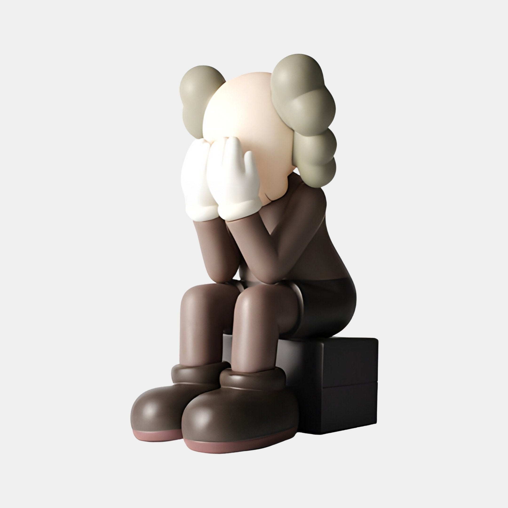 The Hazelnut Brown Iconify Melancholy Sitting Figure by Giant Sculptures is a resin statue depicting a cartoon character with white-gloved hands over its face, featuring a pale head and crossbone-like gray ears, dressed in dark attire and boots, seated on a black block. Height: 28cm.