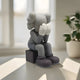 The Slate Grey Iconify Melancholy Sitting Figure by Giant Sculptures is a 28cm cartoonish piece featuring a gray figure with gloved hands on its face, sitting on a black block with large shoes and hair-like shapes. Sunlight highlights the room and a nearby potted plant.