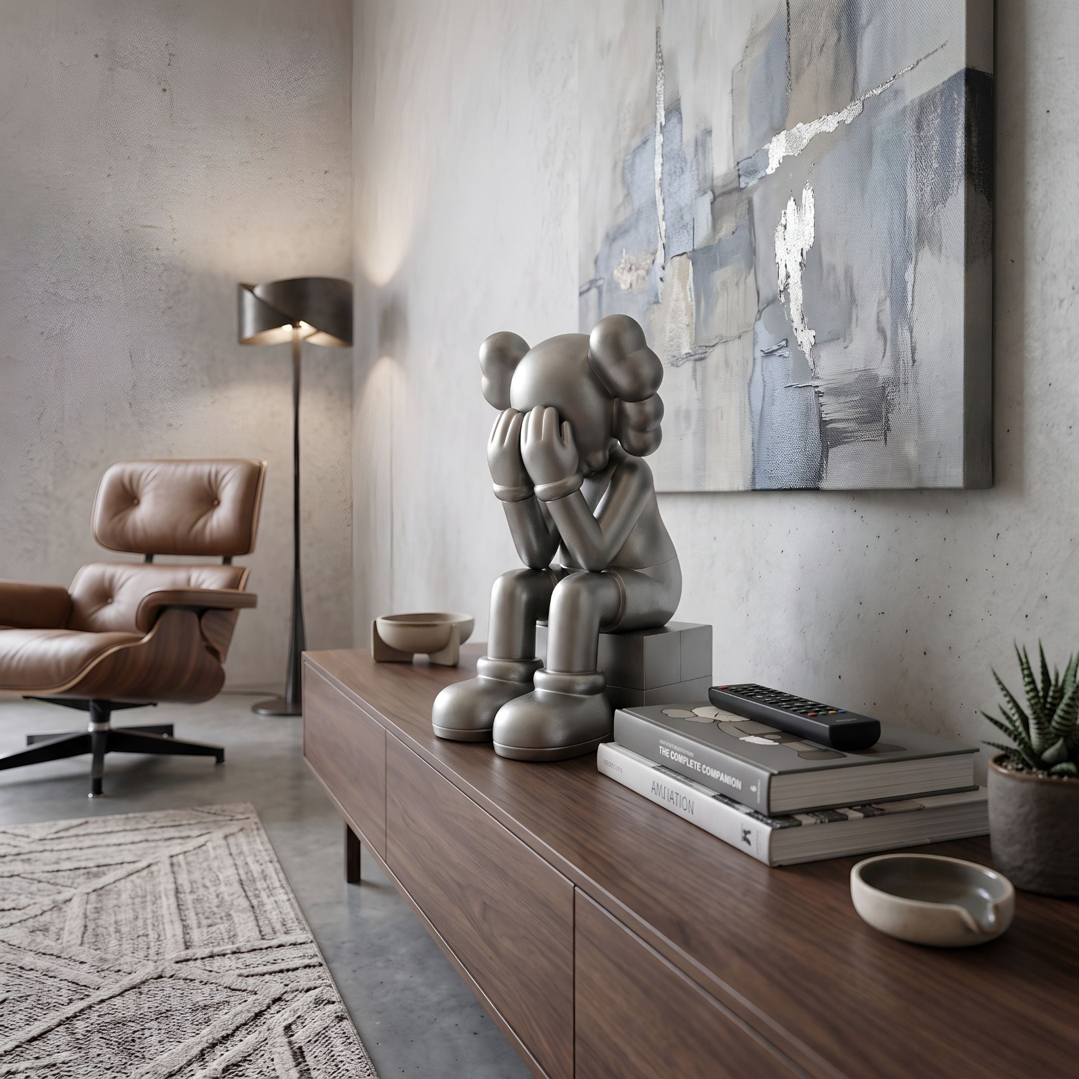 Titanium Slate Iconify Melancholy Sitting Figure, 28cm, in gunmetal finish on a walnut sideboard beside books and a leather lounge chair.
