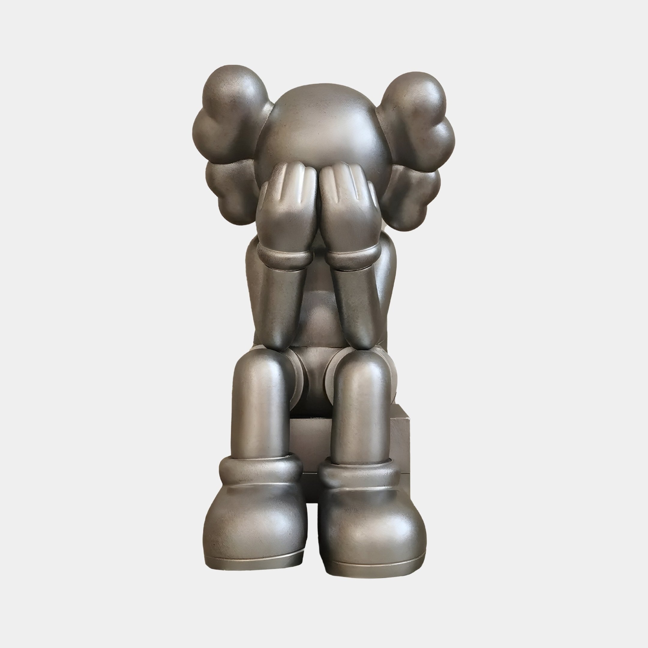 This modern pop-art sculpture by Giant Sculptures, titled Titanium Slate Iconify Melancholy Sitting Figure - 28cm, showcases a cartoonish metallic figure sitting with its face in hands, large rounded shoes, and iconic cloud-shaped ears against a plain background.