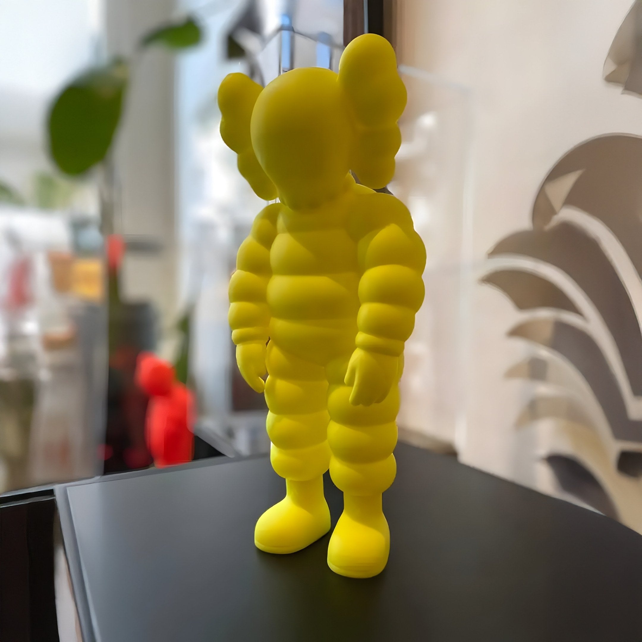 The 28cm Citrus Yellow Iconify Rollo Figure by Giant Sculptures, featuring a contemporary balloon-like body and rounded ears, serves as an artistic centerpiece on a black surface with a subtly blurred backdrop of green and red shades.