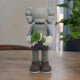 The Soft Grey Iconify Dollars Figure - 30cm by Giant Sculptures is a resin piece with cartoonish limbs and large ears, clutching green bills, symbolizing materialism. A plant in the background enhances the serene setting by the window.