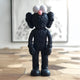 On a wooden floor, the 30cm Obsidian Black Iconify Furry Figure by Giant Sculptures captivates art collectors with its oversized white eyes and pink nose, resembling a whimsical cartoon amid blurred framed artwork.