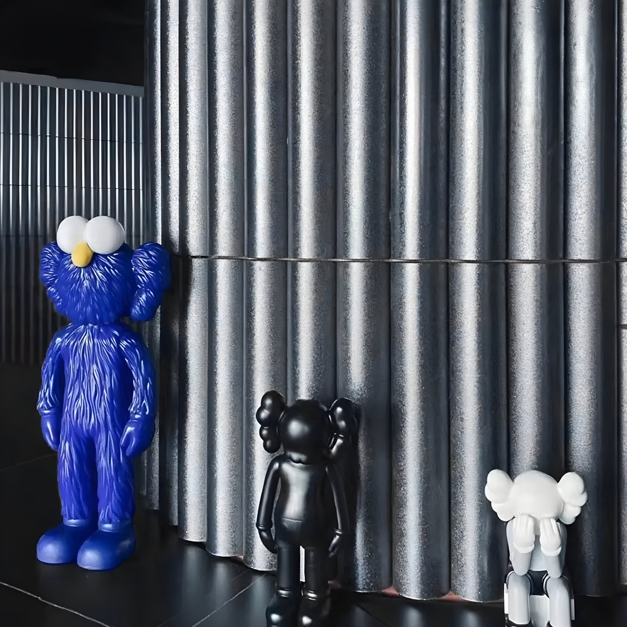 The scene features three cartoon-like statues as embodiments of contemporary art beside large, metallic cylindrical pillars. The 30cm Deep Blue Iconify Furry Figure by Giant Sculptures resembles a friendly monster, while the black and white ones add playful sculptural craftsmanship.