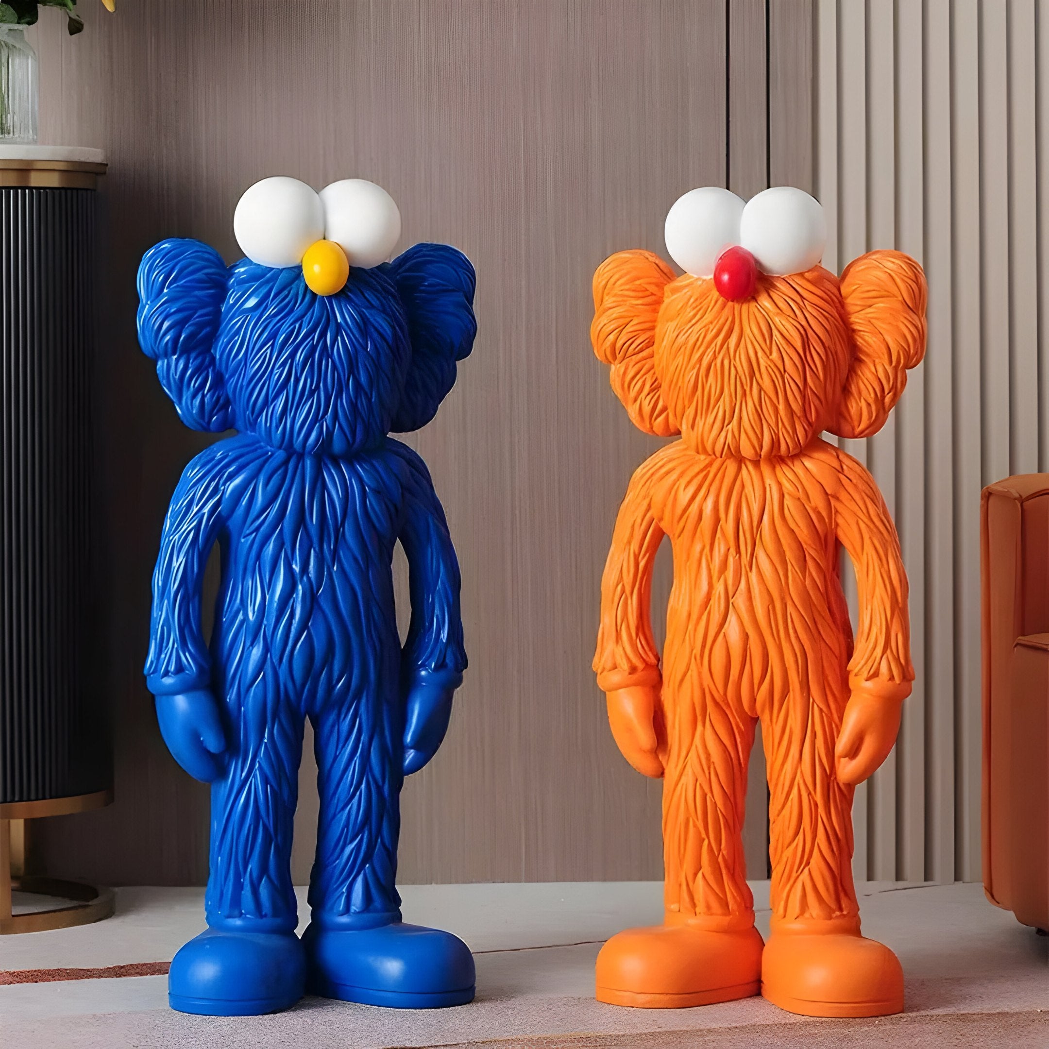 Two vibrant sculptures from Giant Sculptures embody contemporary art, resembling cartoon-like characters: the Deep Blue Iconify Furry Figure - 30cm with a yellow nose and an orange counterpart with a red nose. Both feature large white eyes and fur-like textured surfaces.