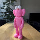 The Petal Pink Iconify Furry Figure by Giant Sculptures, featuring large white eyes and exaggerated ears and nose, captures a playful pop-art aesthetic. It stands charmingly on a wooden table next to a leafy plant and black TV against a light brick wall.